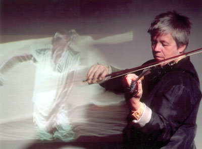 A photo of Steina Vasulka playing her violin in front of a projection on the wall in her piece 